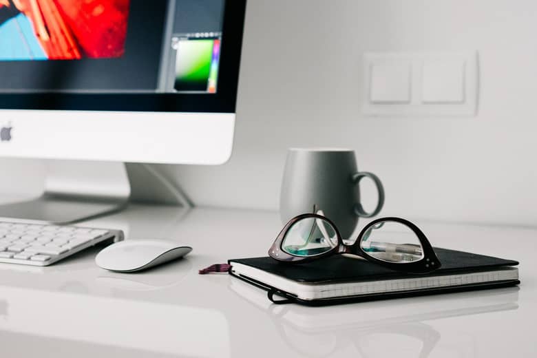 Office desk with Mac, notepad, cup and glasses.