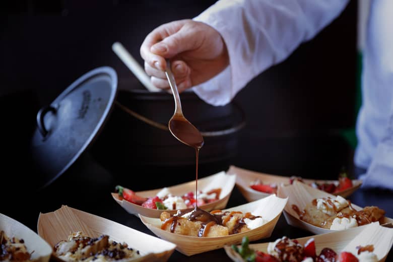 Chef spoon pouring hot chocolate sauce over banana split.